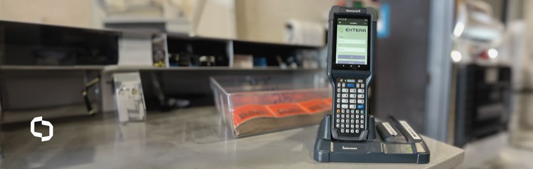 A handheld Honeywell device with asset tracking software displayed on the screen, docked on a counter in an industrial setting. This technology enhances asset management and supply chain visibility by streamlining tracking and logistics operations.