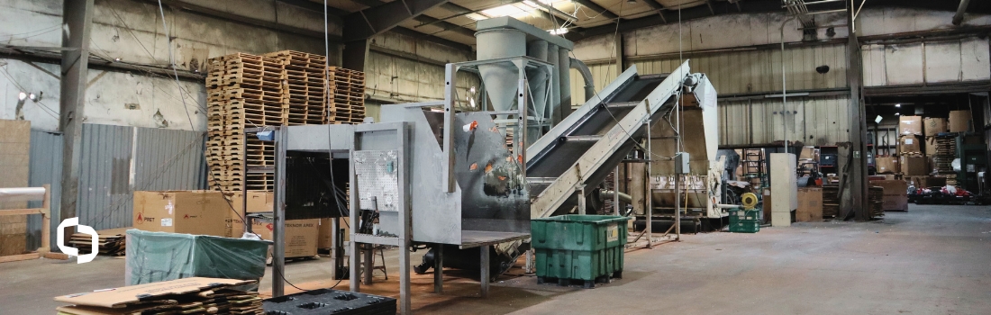 An industrial facility showcasing machinery used in the plastic recycling process, including a conveyor system and shredder surrounded by pallets and storage bins. This setup highlights the scale and efficiency of sustainable manufacturing solutions.
