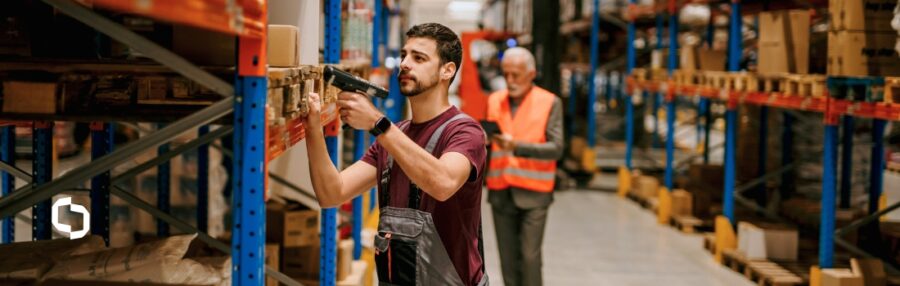 A warehouse worker scans a box on a shelf using a handheld device, while another employee checks inventory in the background. This image illustrates modern supply chain trends such as container tracking and RFID tracking, supporting broader sustainability trends in inventory and logistics management.