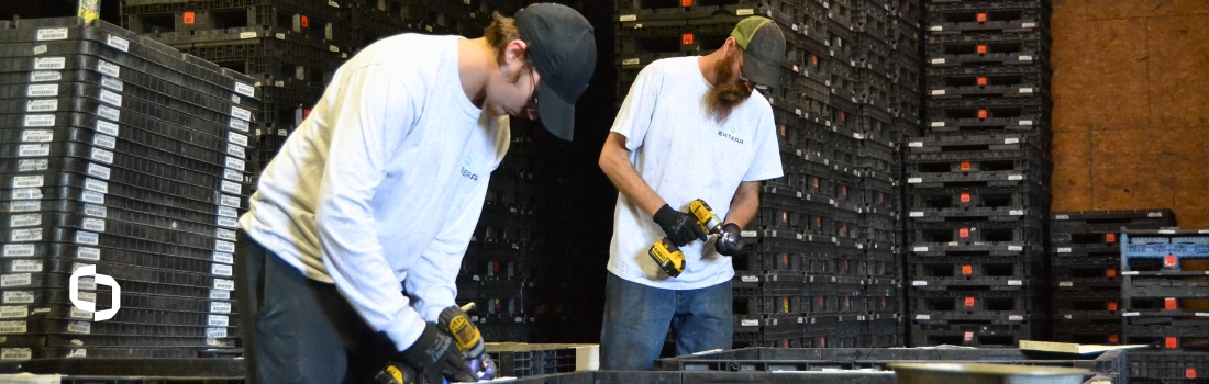 Extera warehouse workers use power drills to assemble reusable packaging containers, with stacks of black plastic containers lining the warehouse behind them.