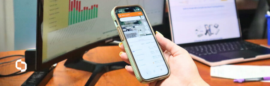 Scoutwise app open on a phone for asset tracking, with supply chain visibility software dashboards on the monitor and laptop behind it