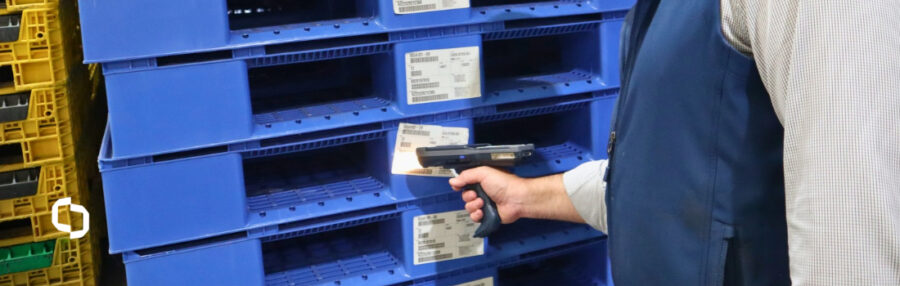 Worker using a Handheld scanner to read barcode asset tracking tags on stacked blue plastic pallets, supporting BLE tracking in a busy warehouse