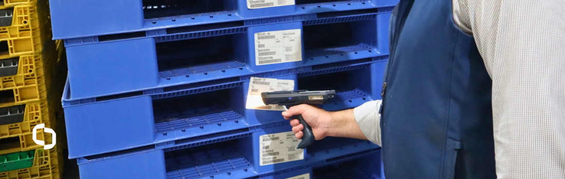 Worker using a Handheld scanner to read barcode asset tracking tags on stacked blue plastic pallets, supporting BLE tracking in a busy warehouse